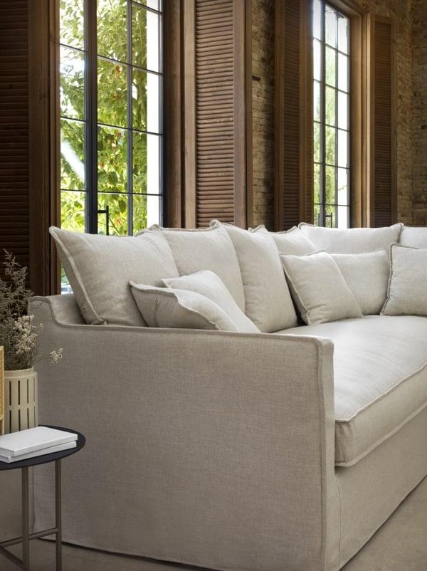 Sofá chaise longue tapizado en blanco roto, estilo rústico moderno con fondo de piedra natural. Ideal para espacios serenos y luminosos.